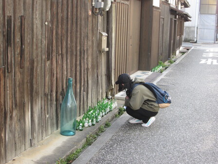 参加者の一人が、建物の壁沿いに置いてある多数の小さな酒瓶を屈んで見ている。その中に容量二升半と思しき大きな瓶が一本ある。